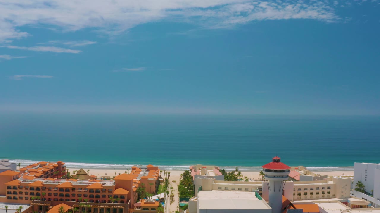 Palmilla Beach aerial frame showing orange and cream beachfront buildings, a white hotel wing, the pale beach, blue ocean, thin clouds, and no visible people.