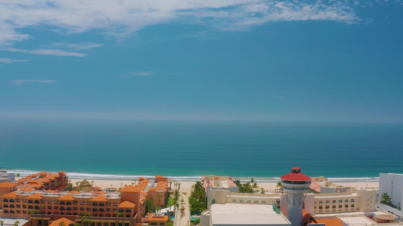 Mid-clip Palmilla Beach view with the red-capped round tower near the lower right, orange-roofed buildings to the left, white hotel structures, calm ocean, and no visible people.