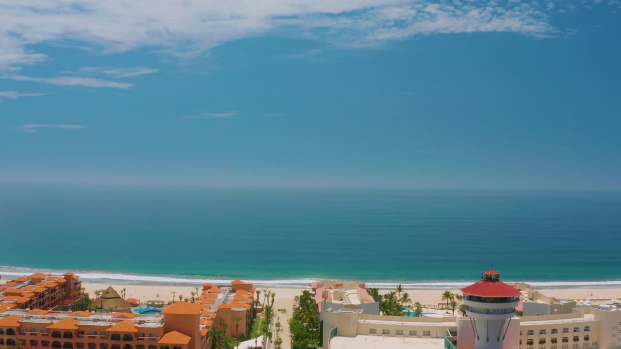 Wide aerial frame over Palmilla Beach, Baja California Sur, with orange-roofed buildings, a red-roofed round structure, pale sand, calm blue water, clear sky, and no visible people.