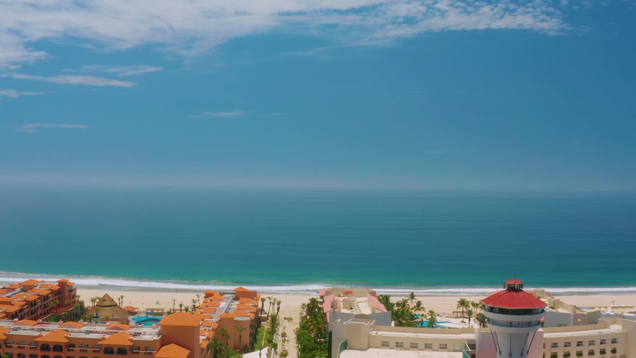 High aerial frame over Palmilla Beach with orange resort buildings at left, palms and pools near the beach, open turquoise water beyond the sand, and no visible people.