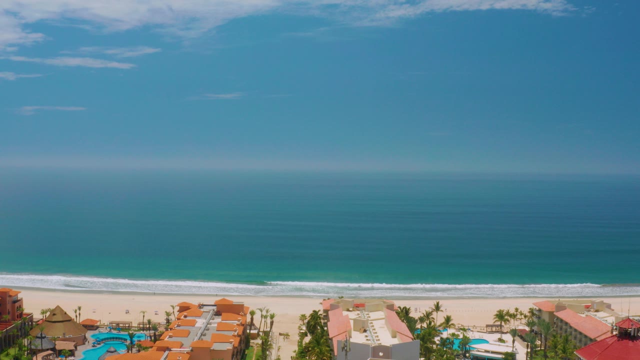 Opening frame over Palmilla Beach, Baja California Sur, showing a level ocean horizon, turquoise water, white surf, pale sand, terracotta roofs, and no visible people.
