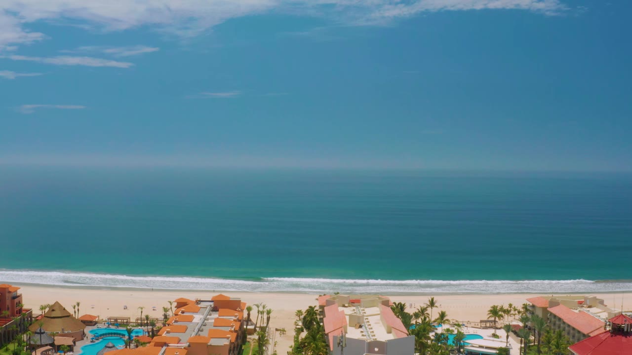 Aerial beachfront view with calm ocean, surf line, sand, roofs, palms, and pools.