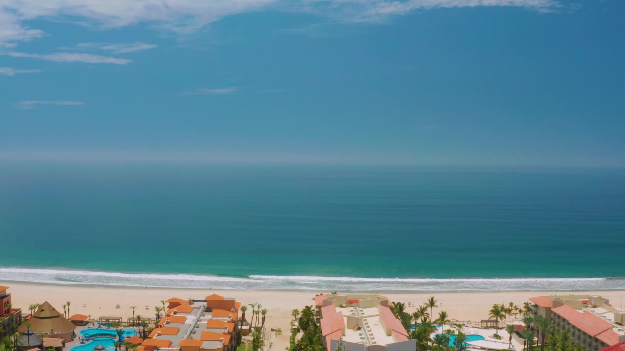 A high view across Palmilla Beach shows deep blue water fading to turquoise near shore, a thin white surf band, sand, palms, rooftops, and no visible people.