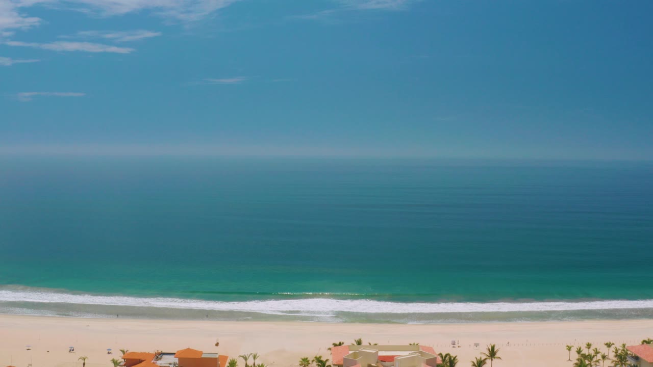 Opening frame of Palmilla Beach in Baja California Sur with layered blue sea, a white wave line, pale sand, small palms and orange roofs at the bottom edge, and no visible people.