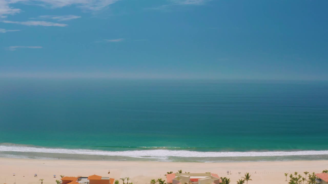 Palmilla Beach frame with the ocean filling most of the view, thin clouds near the top, a bright surf band near the sand, low orange-roofed buildings at the bottom edge, and no visible people.