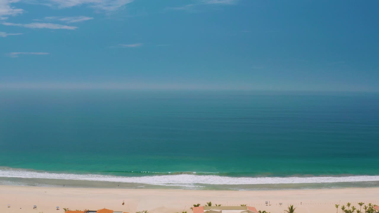Palmilla Beach view at 0:02 with a steady blue horizon, mild waves rolling toward pale sand, faint clouds above, small palms and rooflines at the lower border, and no visible people.