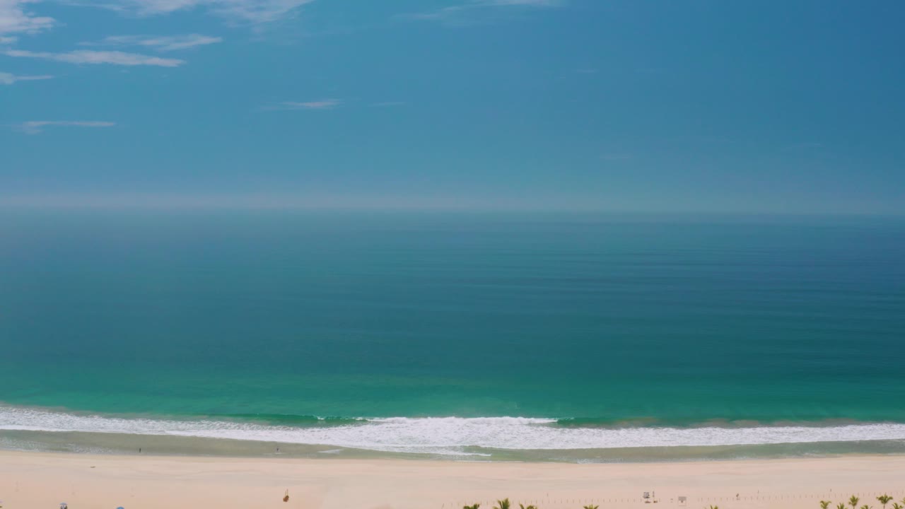 Ocean and beach view with teal water, white breaking waves, pale sand, and a few tiny beach marks.