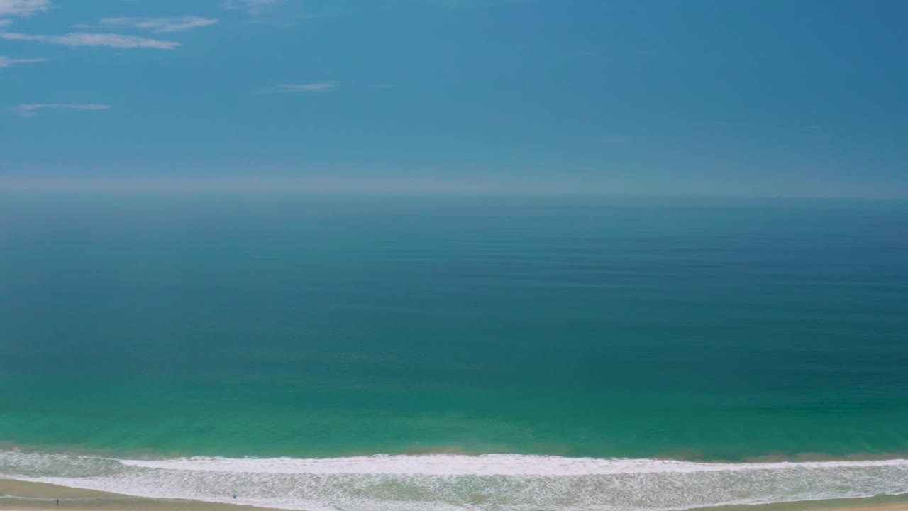 Elevated Palmilla Beach frame showing a level horizon, faint clouds in the blue sky, broad turquoise water, white surf, a sliver of sand, and no visible people.