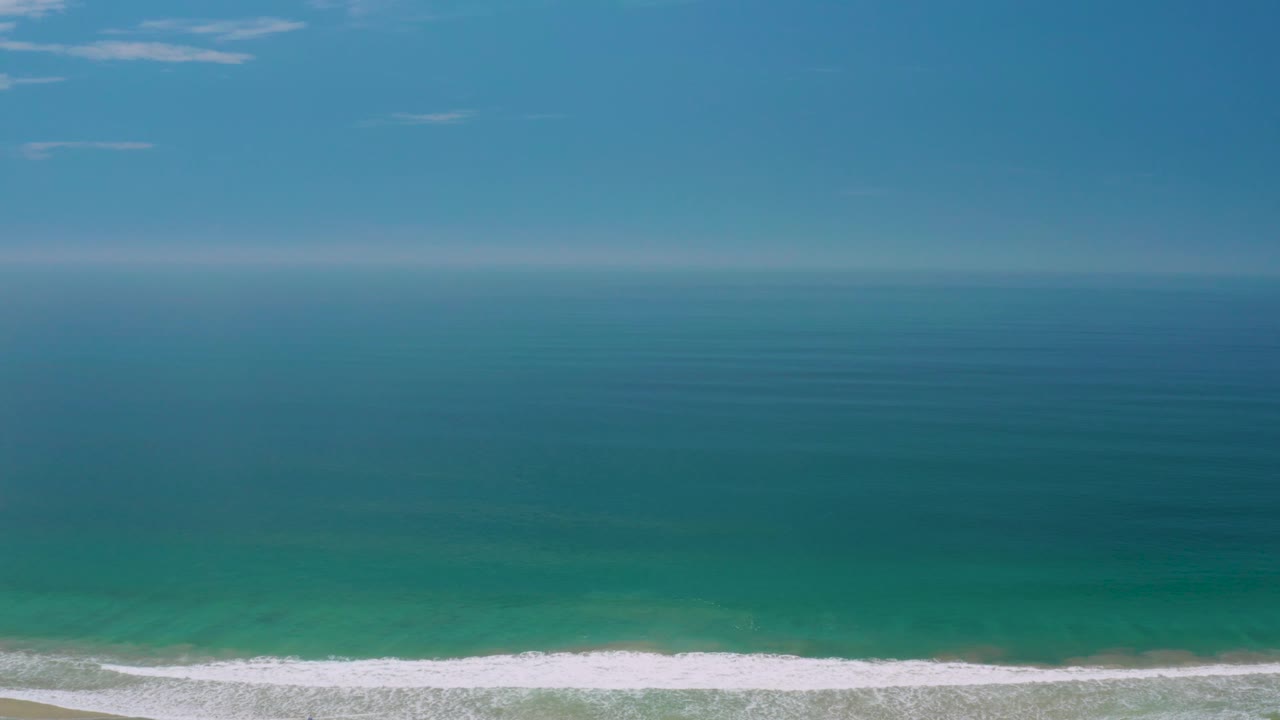 Palmilla Beach water fills most of the frame, with blue sky above, a hazy horizon, darker offshore water, pale green shallows, white surf, and no visible people.