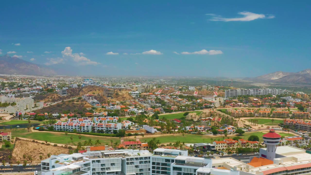 Opening aerial frame over Palmilla Beach, Baja California Sur with white buildings in the foreground, green grounds and dry hills in the middle distance, mountains on the horizon, and no visible people.