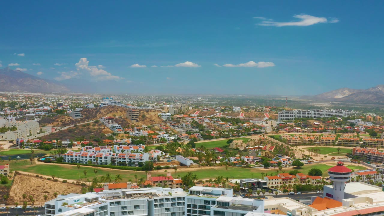 At 0:02, the Palmilla Beach aerial frame shows resort-style rooftops at the bottom, a dry hill with buildings near center, a hazy urban background, mountains, and no visible people.