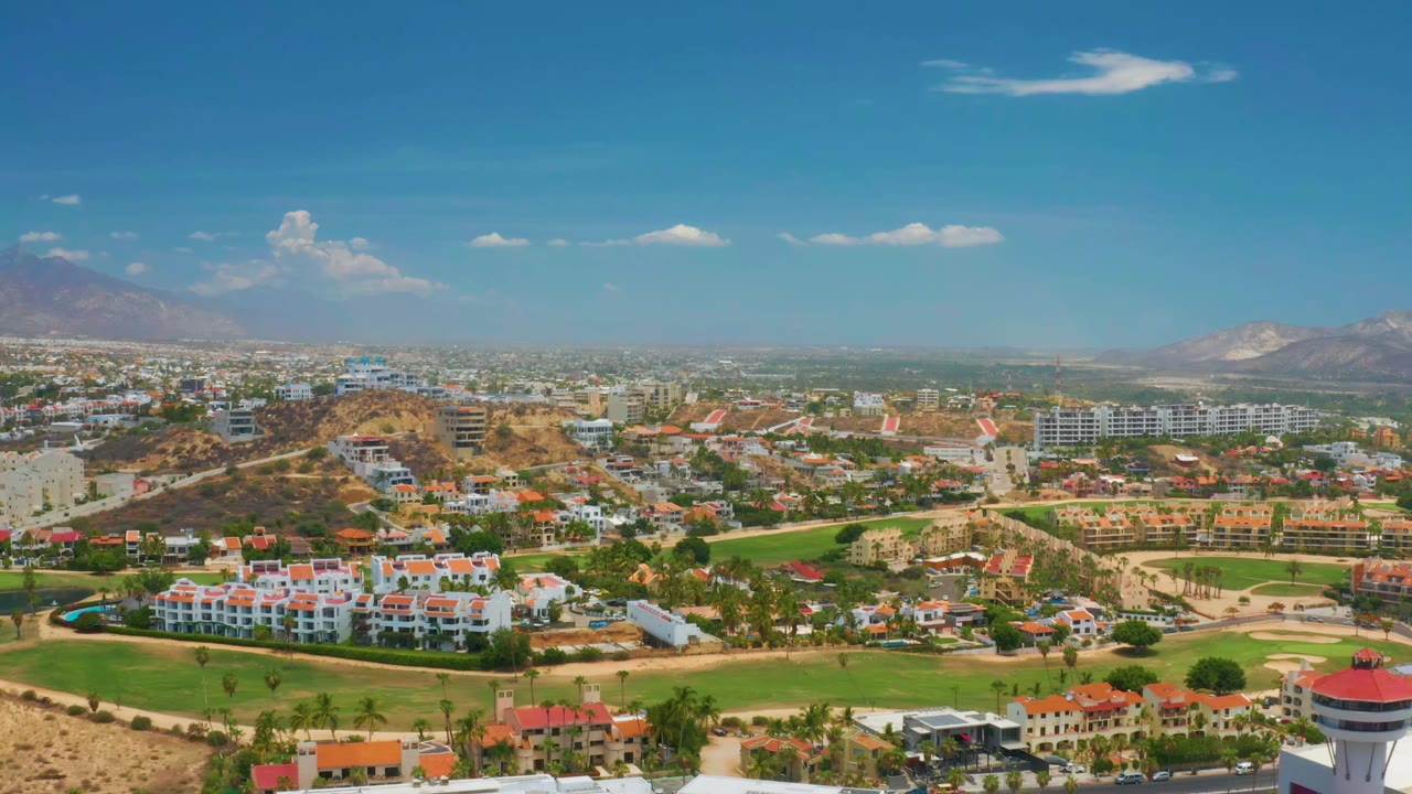 Elevated Palmilla Beach view with white resort buildings in the foreground, green fields and roads through the middle ground, hazy distant development, mountains, and no visible people.