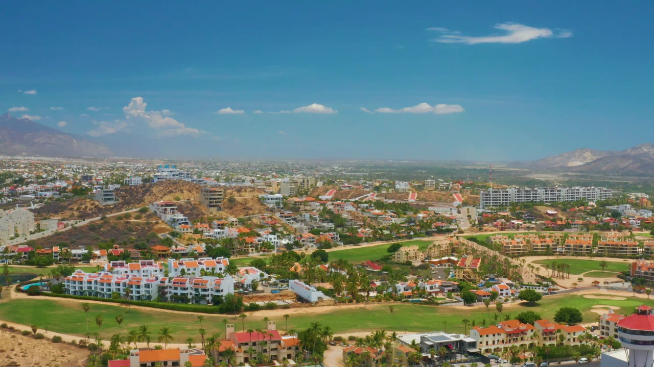 High Palmilla Beach frame showing hotel rows and palms at the lower edge, a dry central hill, surrounding roads and buildings, distant mountains, and no visible people.