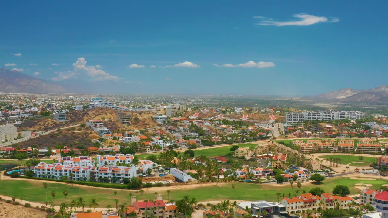 Wide daytime view of resort buildings, green fields, dry hills, and mountains.
