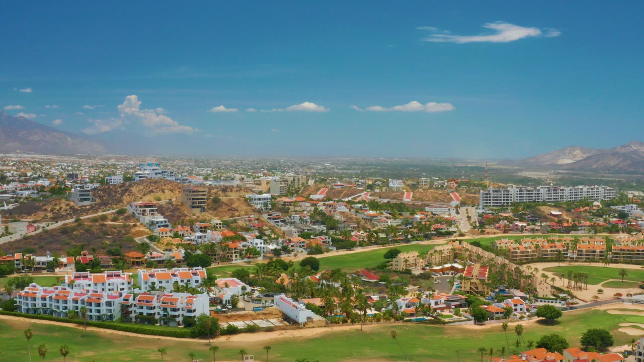 Opening aerial frame over Palmilla Beach, Baja California Sur, showing white buildings, red roofs, green lawns, dry hills, distant mountains, and no visible people.
