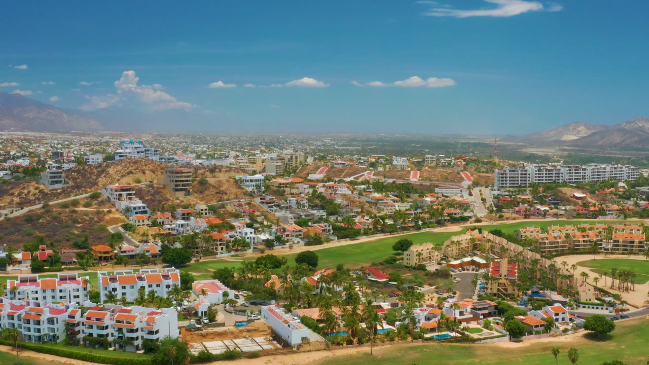 Opening aerial frame over Palmilla Beach, Baja California Sur, with red-roofed white buildings in the foreground, green fairways, dry hills, distant mountains, blue sky, and no visible people.