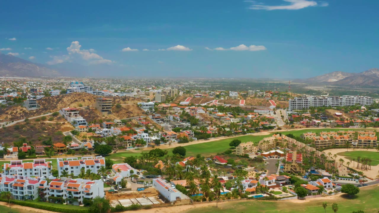 One second into the Palmilla Beach aerial, the view centers on hillside development, landscaped green areas, roads, a broad city horizon, distant mountains, and no visible people.