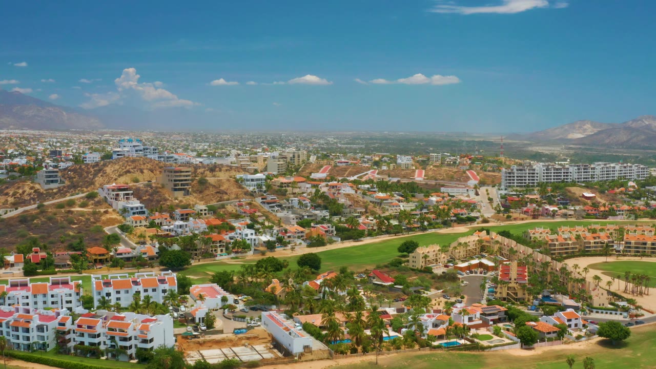 At 0:02, the Palmilla Beach view keeps red-roofed buildings low in frame, tan hills and roads across the middle, a dense distant city area, mountains, and no visible people.