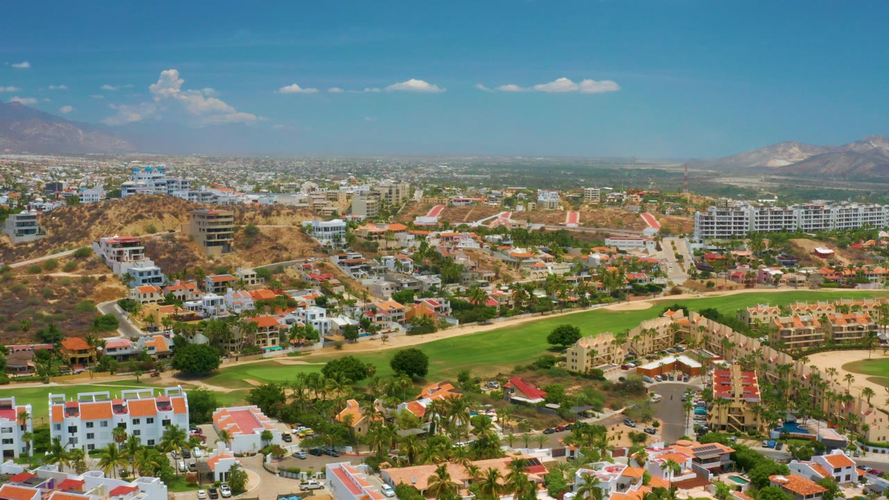 Opening aerial frame over Palmilla Beach, Baja California Sur, showing red-roofed buildings, palm-lined streets, a green fairway, dry hills, distant mountains, and no visible people.