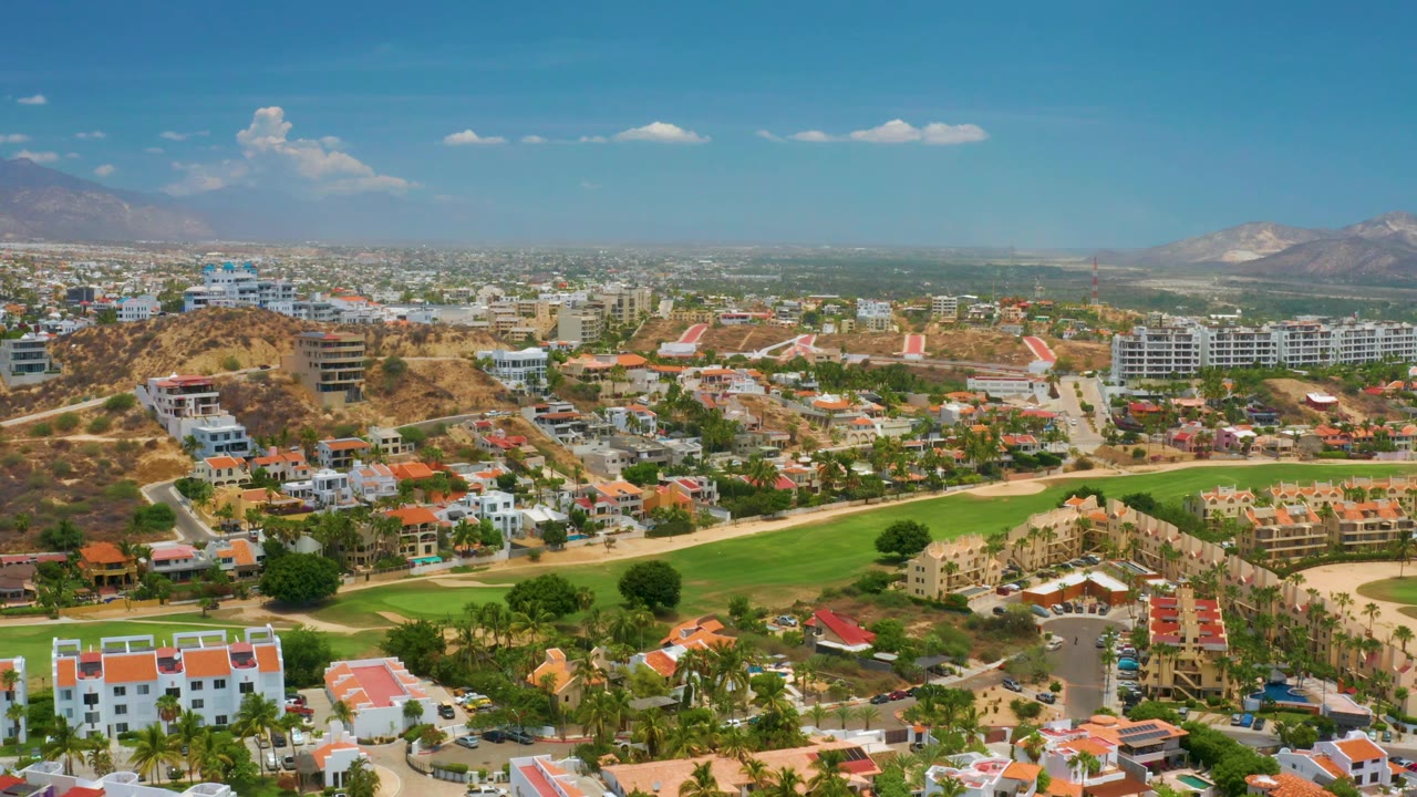 Third aerial frame in Palmilla Beach, Baja California Sur, looking over orange-roofed residences, a long green fairway, dry hills, distant city blocks, mountains, and no visible people.