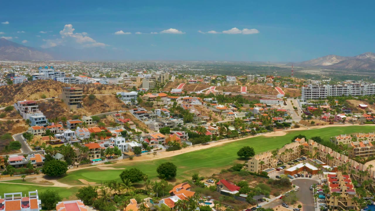 One second into the clip, the aerial view over Palmilla Beach shows the fairway across the lower frame, clustered white buildings, a hazy mountain horizon, and no visible people.
