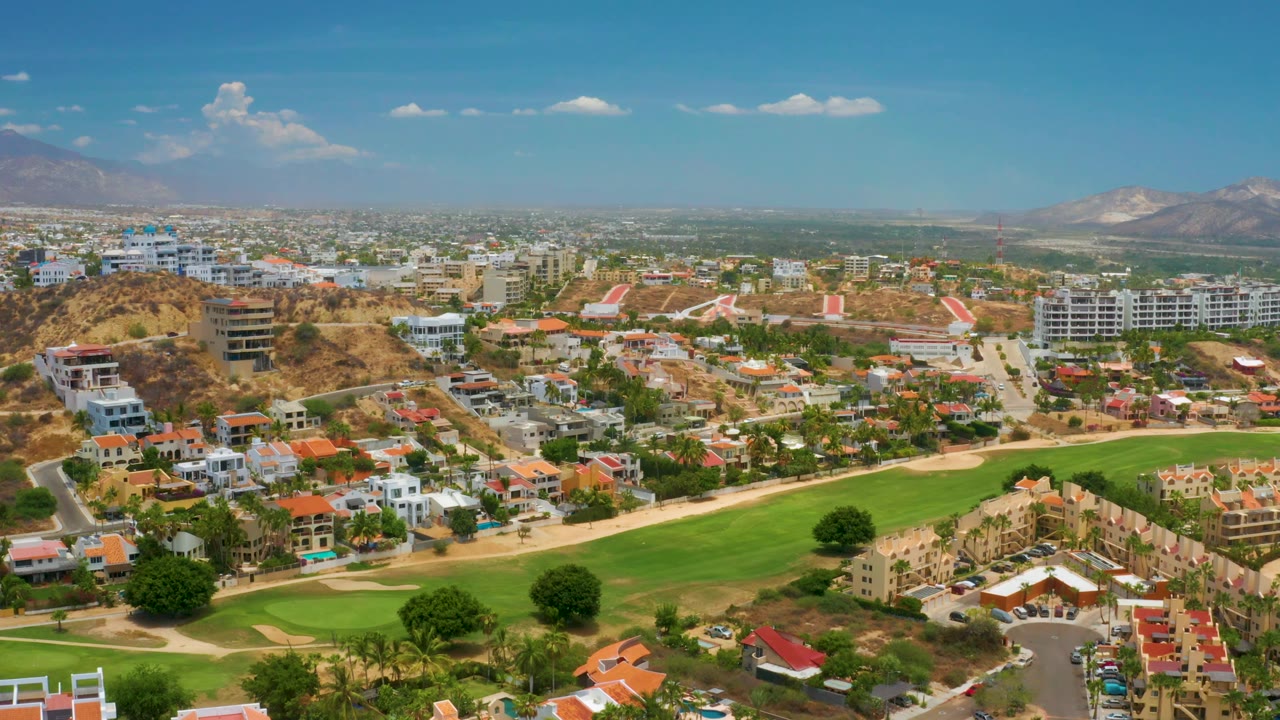 At 0:02, the Palmilla Beach aerial frames a bright green fairway below, terracotta-roofed homes and white buildings in the middle distance, dry hills, mountains, and no visible people.