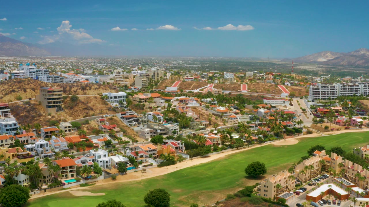 Aerial view over Palmilla Beach, Baja California Sur, with a green golf fairway in the foreground, villas and condos beyond it, distant mountains, and no visible people.