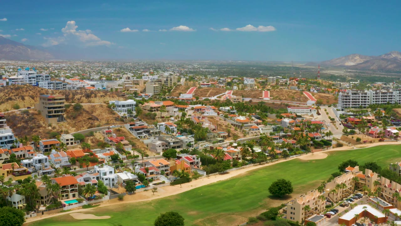 Aerial view across Palmilla Beach with villas and condos surrounding a green fairway, palm-lined streets, hazy mountains in the background, and no visible people.