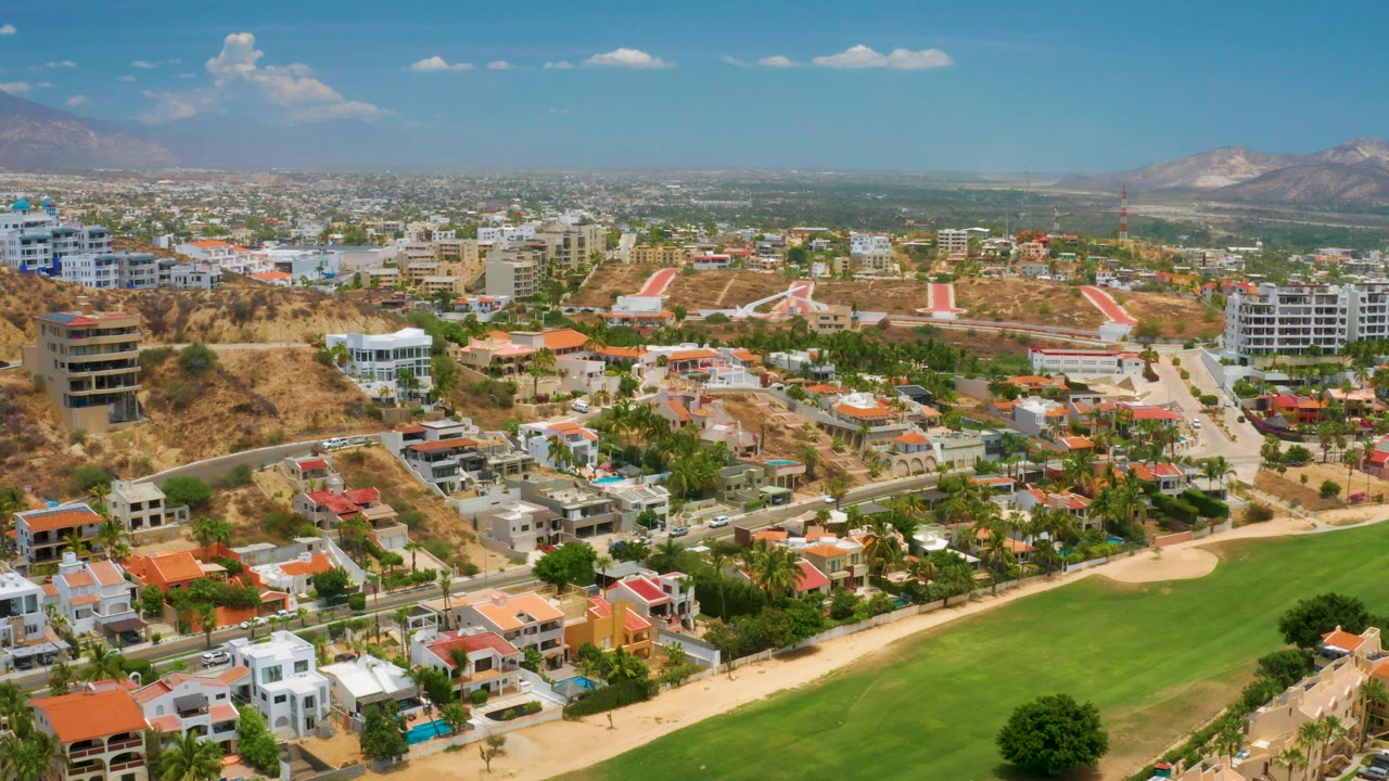 Aerial frame over Palmilla Beach, Baja California Sur showing colorful houses, dry hills, a green foreground field, distant mountains, blue sky, and no visible people.