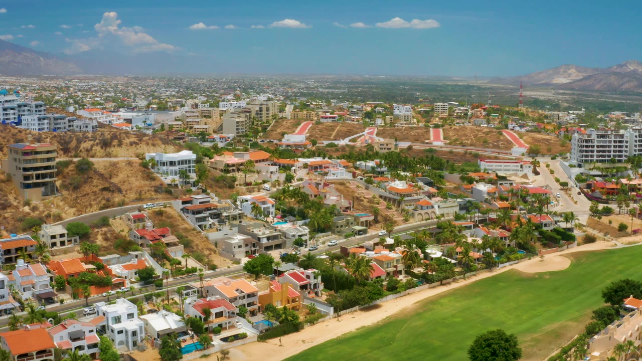 Aerial view in Palmilla Beach, Baja California Sur with villas and low-rise buildings spread across dry hills, a bright green field at lower right, mountains, and no visible people.