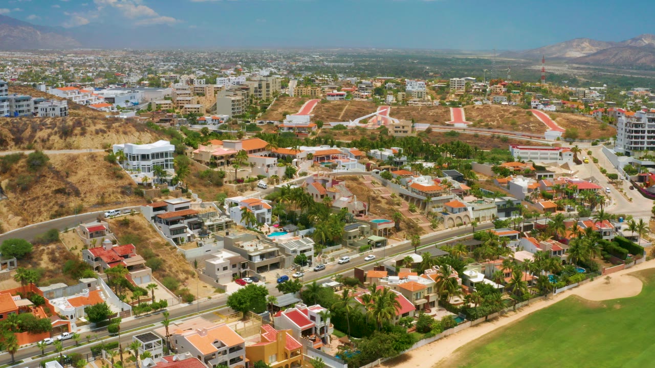 Wide aerial frame over Palmilla Beach, Baja California Sur, showing hillside homes, curving roads, a green landscaped area, dry slopes, distant mountains, blue sky, and no visible people.