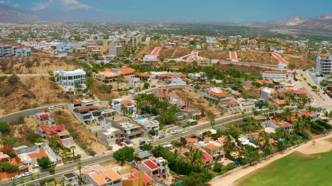 Palmilla Beach aerial panorama showing a green course or landscaped field at the bottom edge, residential roofs, hillside roads, a brown ridge, hazy mountains, and no visible people.