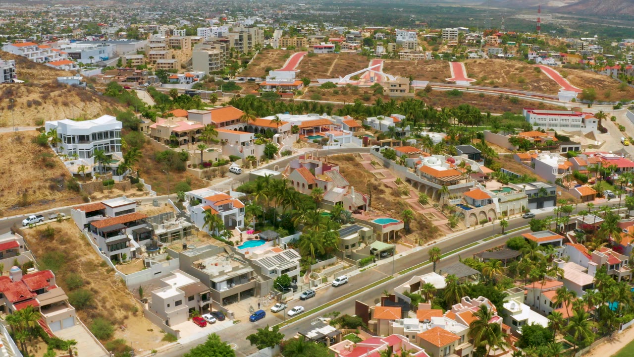 Palmilla Beach aerial frame showing large homes with orange tile and flat roofs, private pools, dry grassless yards, palm trees, paved roads, distant buildings, and no visible people.