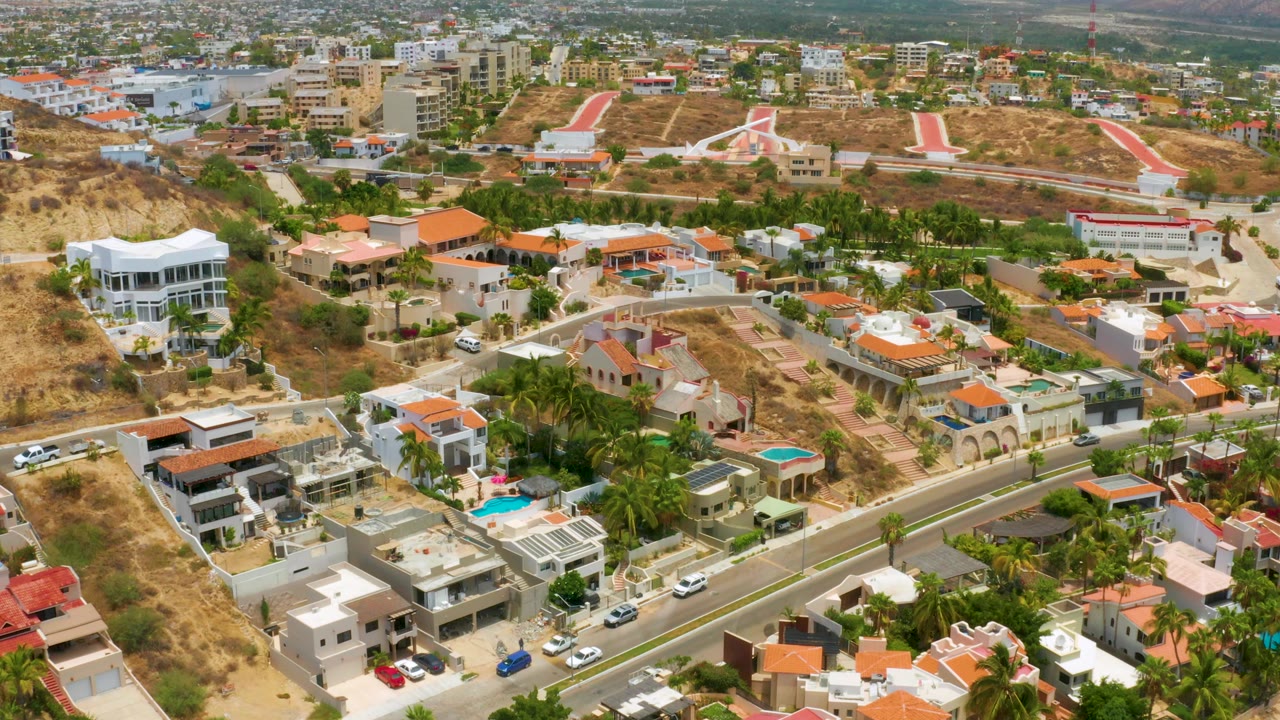 Palmilla Beach frame looking over villas with private pools, palm-lined yards, dry earth lots, a main paved road, distant low-rise buildings, and no visible people.