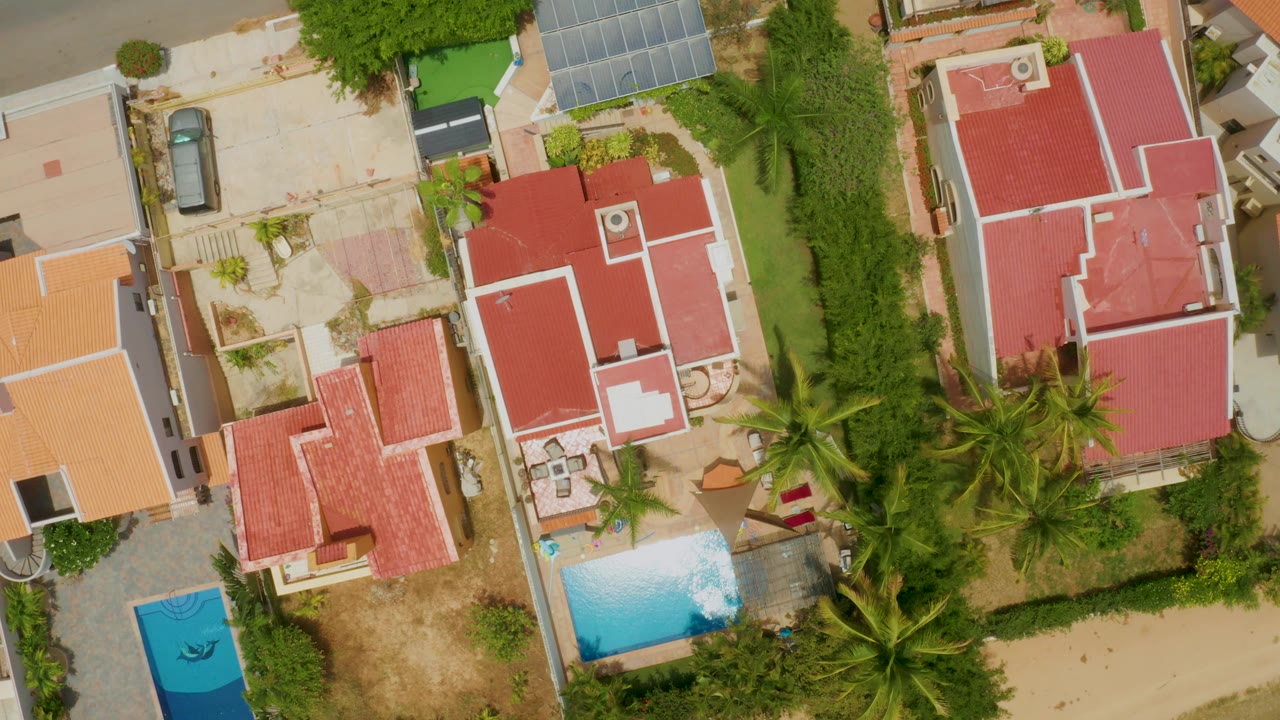 Palmilla Beach overhead frame showing residential roofs, a turquoise pool, palms along the property edge, pale patios, dry landscaping, and no visible people.