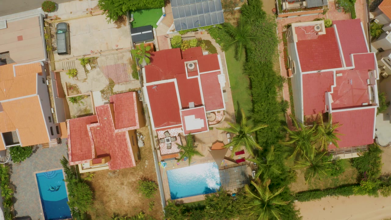 Straight-down view of tiled roofs, a blue pool, palms, and adjacent lots.