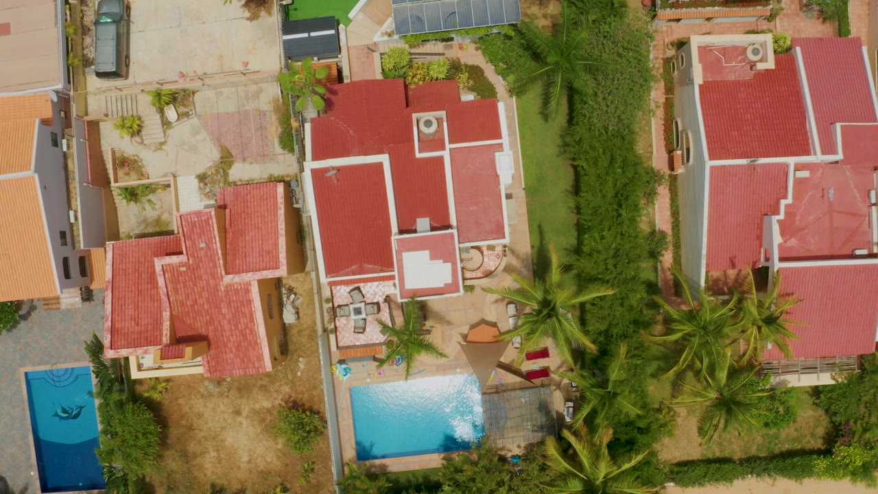 Overhead Palmilla Beach frame with the central red-roofed house larger in view, a pool corner near the bottom, dense palms to the right, and no visible people.