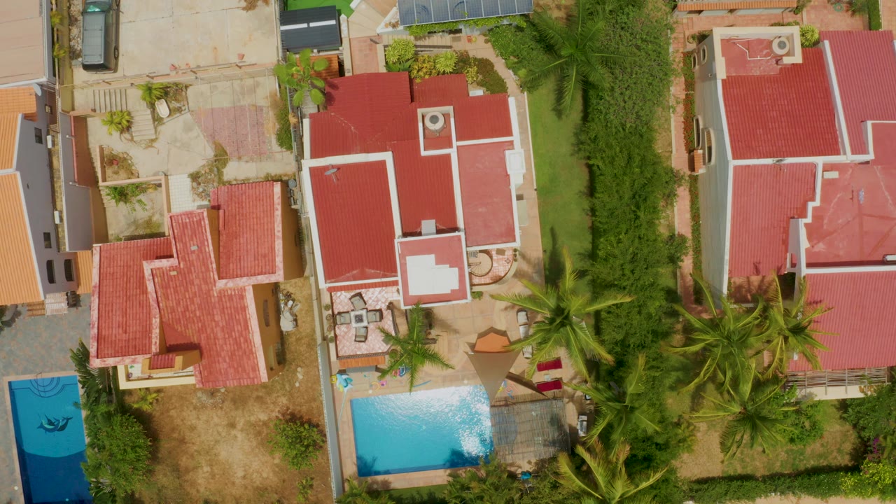 A Palmilla Beach residential aerial frame showing red roofs, a bright rectangular pool near the lower left, palms across the lower right, and no visible people.