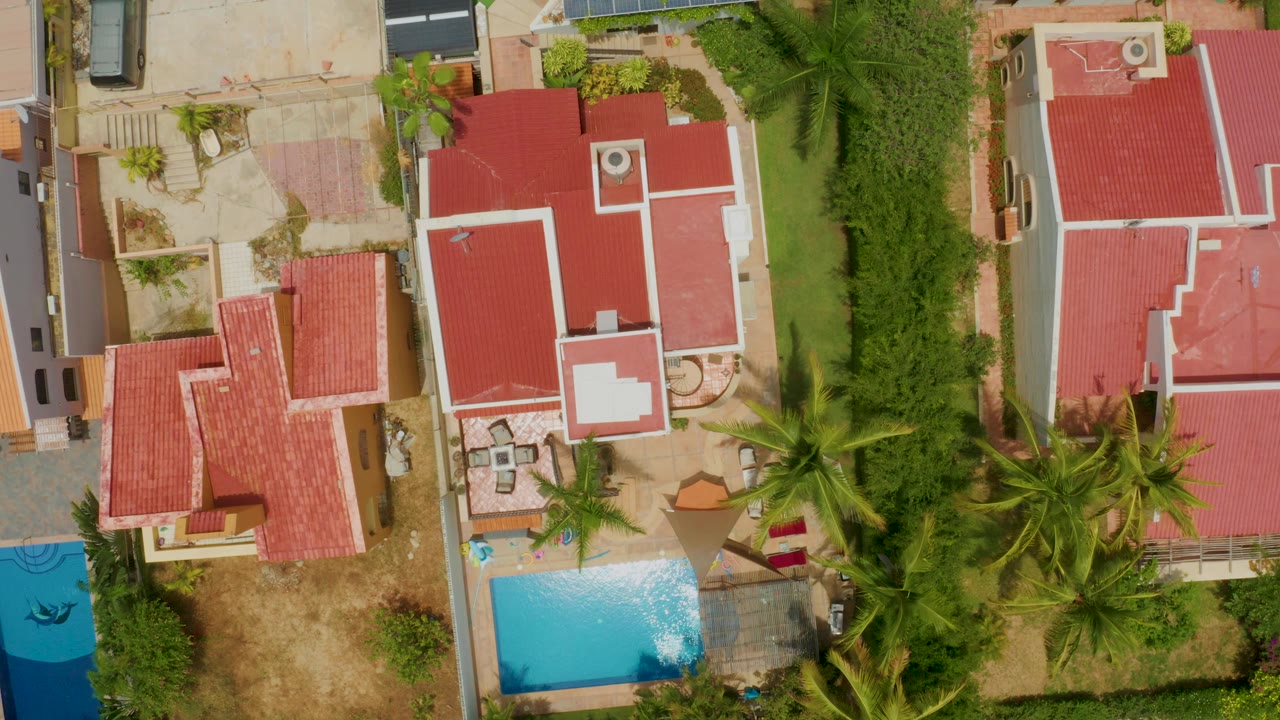 Overhead residential view with red roofs, a pool, palms, and a blue vehicle or cover at left.