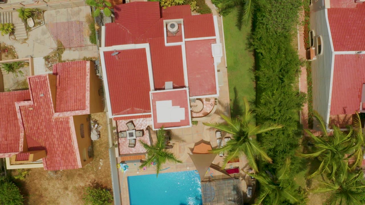 Overhead aerial frame in Palmilla Beach, Baja California Sur showing red-roofed residences, a turquoise pool at lower left, palms, hedges, patio areas, and no visible people.