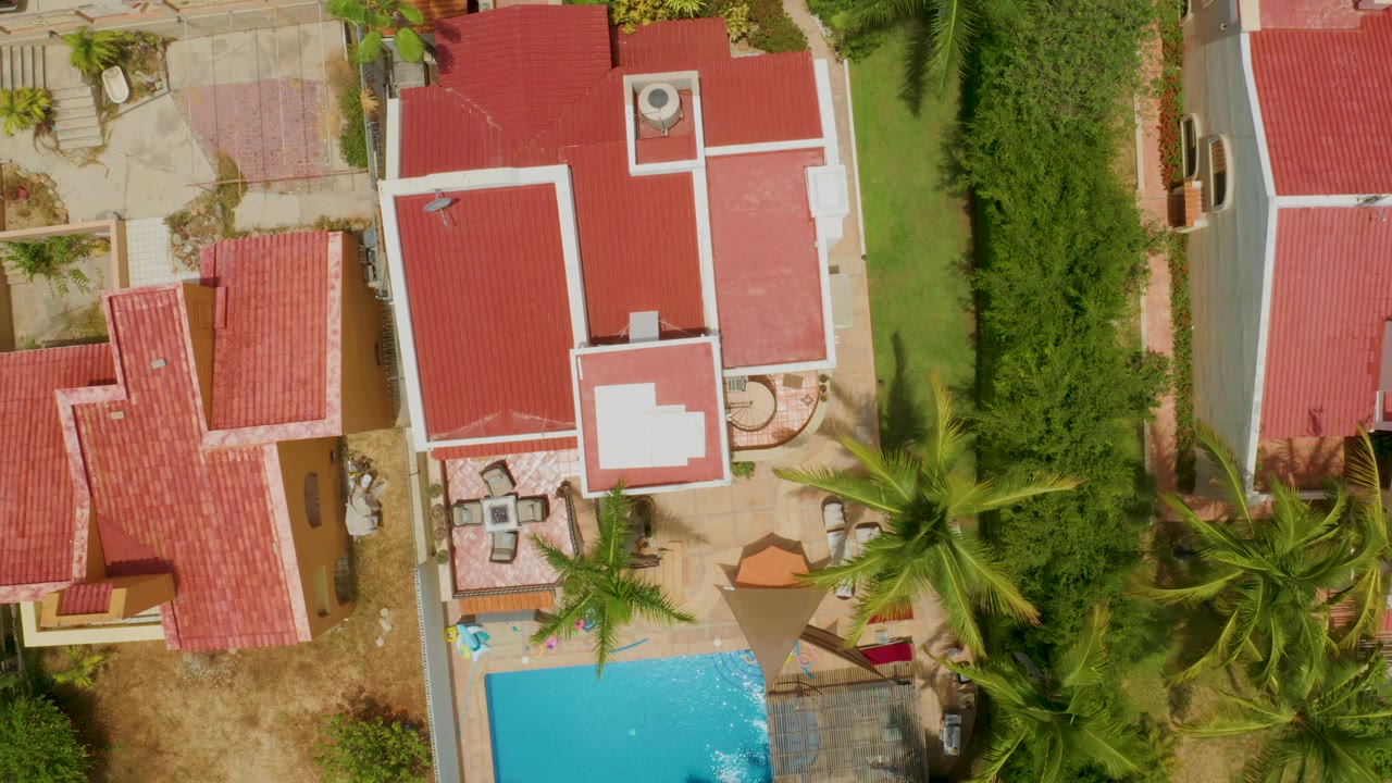 Top-down drone view of a red-roofed house beside a pool and palms.