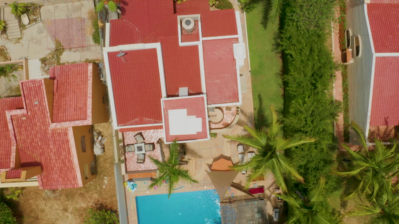 Aerial frame over Palmilla Beach residential roofs with bright pool water at bottom, patio furniture near the center-left, palms over the yard, dense hedging to the right, and no visible people.