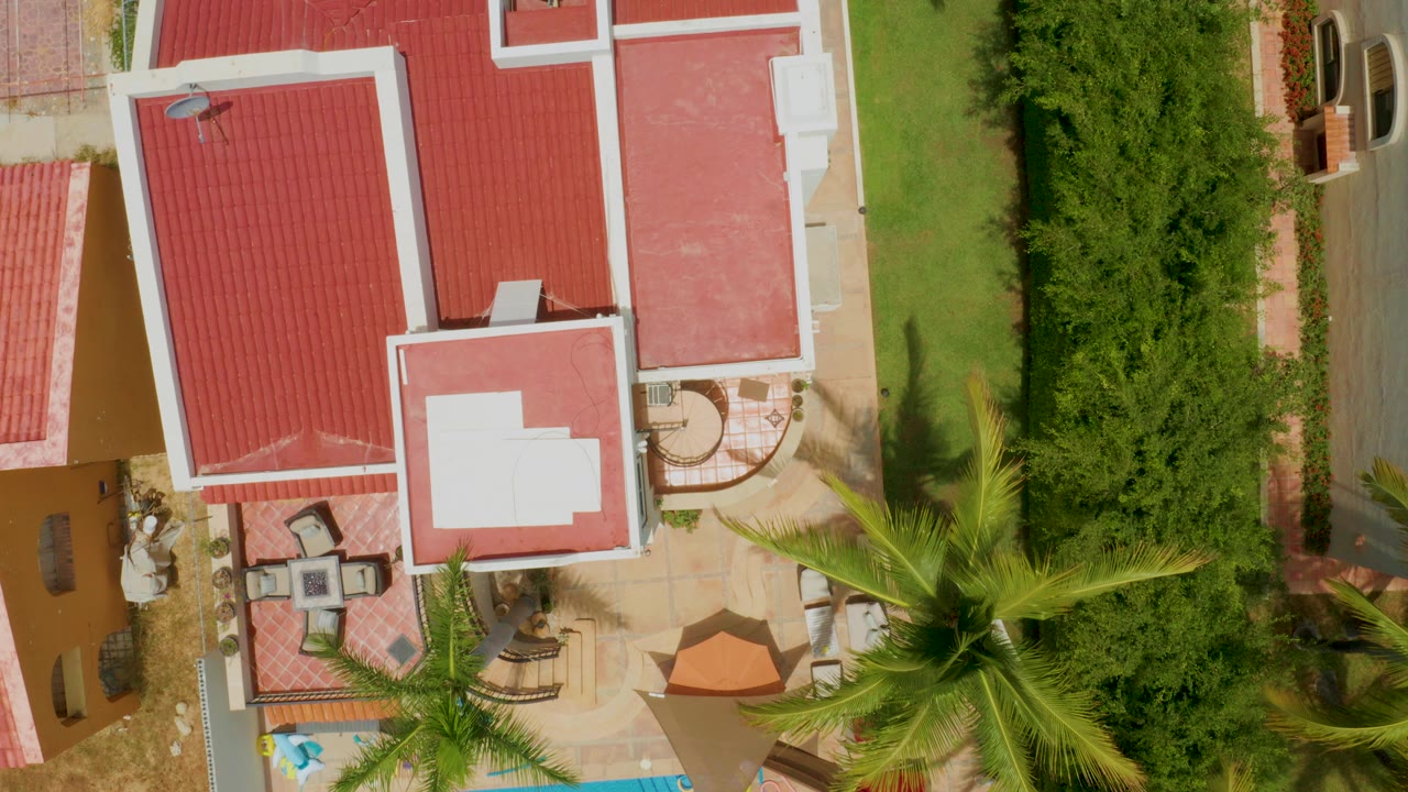 Top-down aerial frame in Palmilla Beach, Baja California Sur showing red roof sections, white roof trim, patio furniture, lawn, hedges, palm fronds, and no visible people.