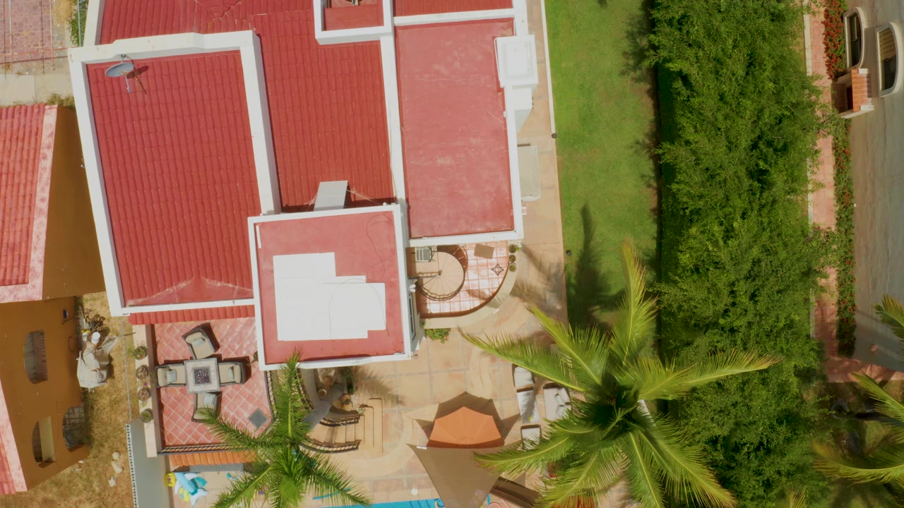 Palmilla Beach aerial frame with the red roof and white parapet centered more tightly, a paved patio below, a palm crown crossing the lower right, and no visible people.