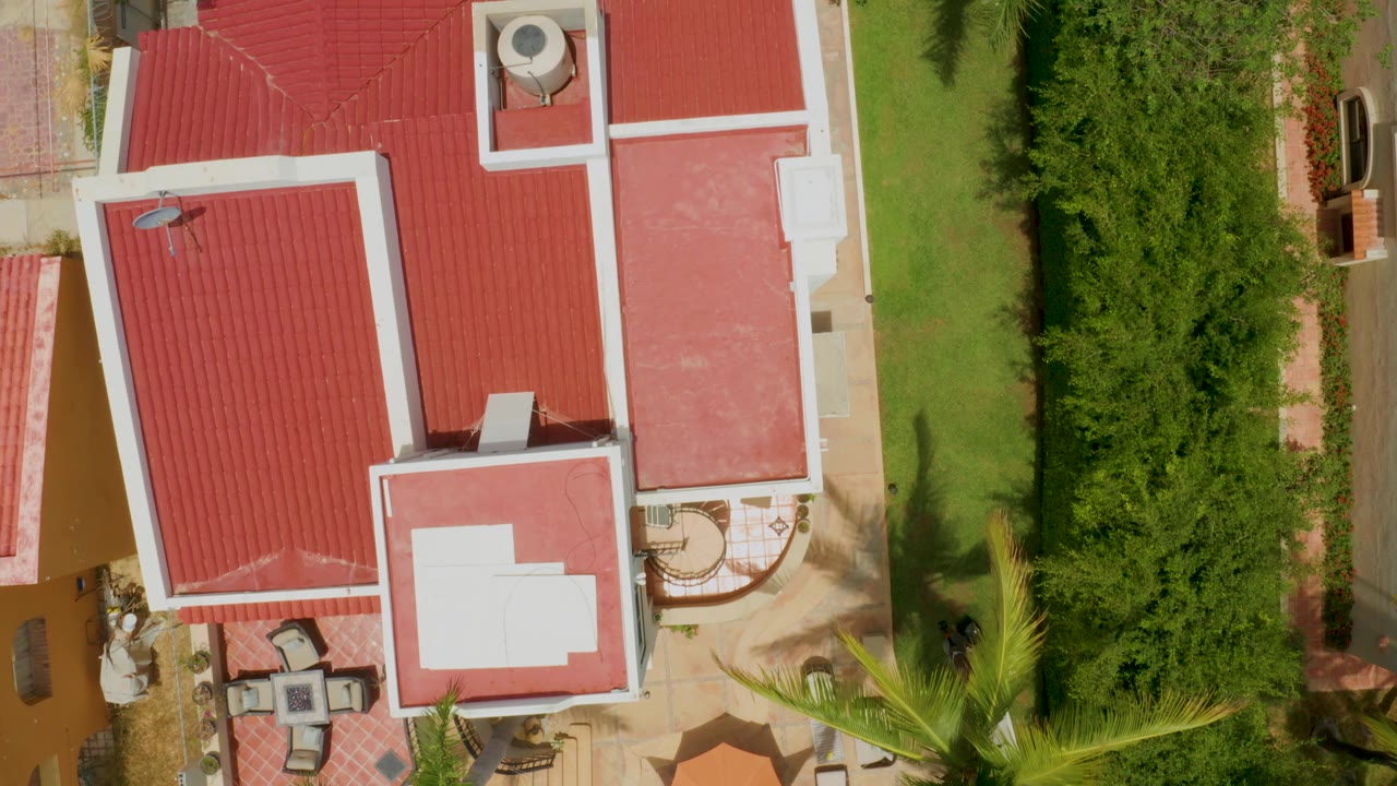 Top-down view in Palmilla Beach with red roof planes, a small rooftop unit, a satellite dish, manicured grass, a dense hedge strip, and no visible people.