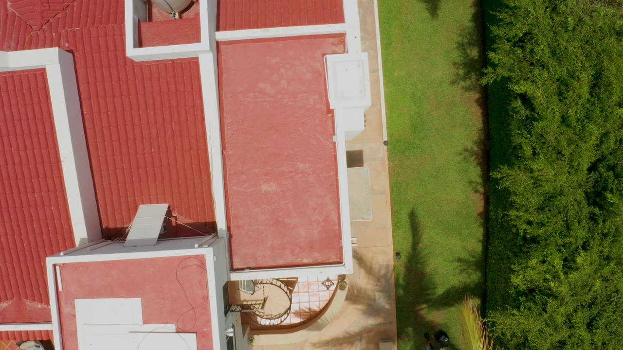 Overhead frame in Palmilla Beach, Baja California Sur showing red sloped and flat rooftops, white trim, a narrow green lawn, dense shrubs, and no visible people.