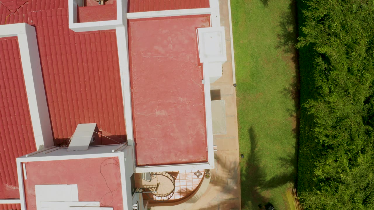 Aerial Palmilla Beach frame showing more of the red sloped roof at left, a flat red roof at center, bright lawn and shrubs at right, and no visible people.