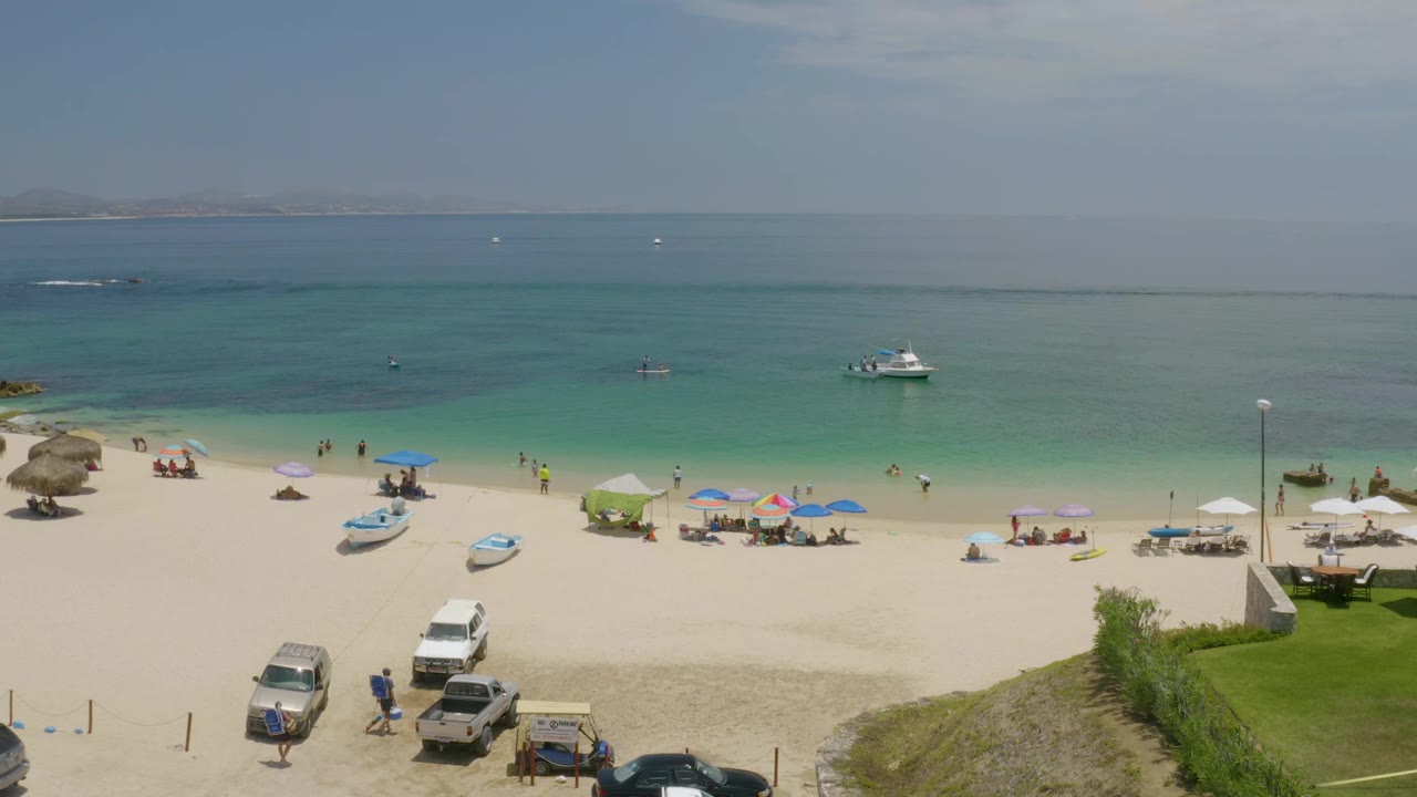 Opening frame of Palmilla Beach in Baja California Sur, Mexico, showing beach umbrellas, small groups of people, parked vehicles, boats on sand, turquoise water, and distant land.
