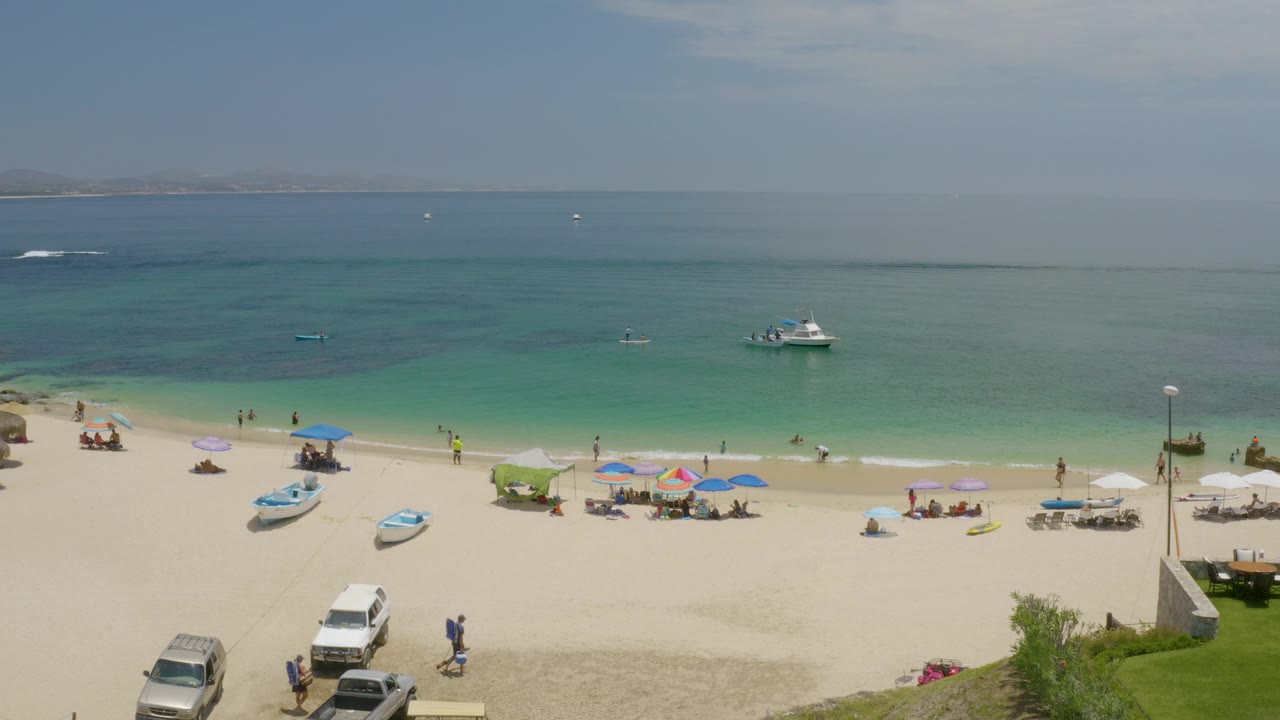 Palmilla Beach, Baja California Sur, shown from above with small boats on the sand and water, scattered umbrellas, beachgoers, and a hazy horizon.