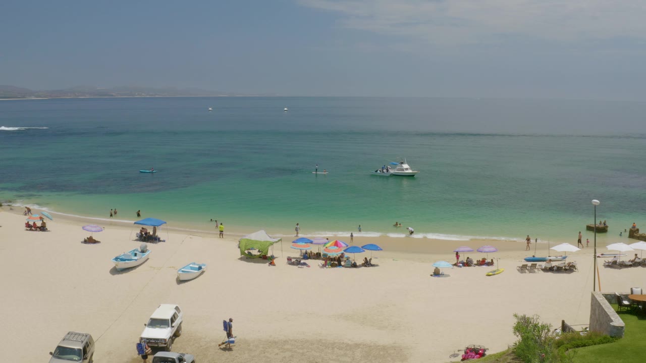 Palmilla Beach is seen from an elevated angle with beachgoers under umbrellas, a lamp post near the shoreline, small boats, and calm water stretching to distant land.